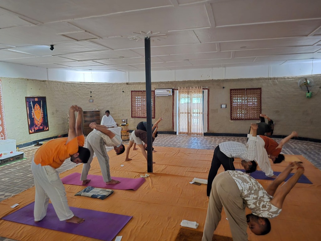 Students practicing Hatha Yoga forward bends in natural mud-walled hall at Adiyog Yogashala with Linga Bhairavi image