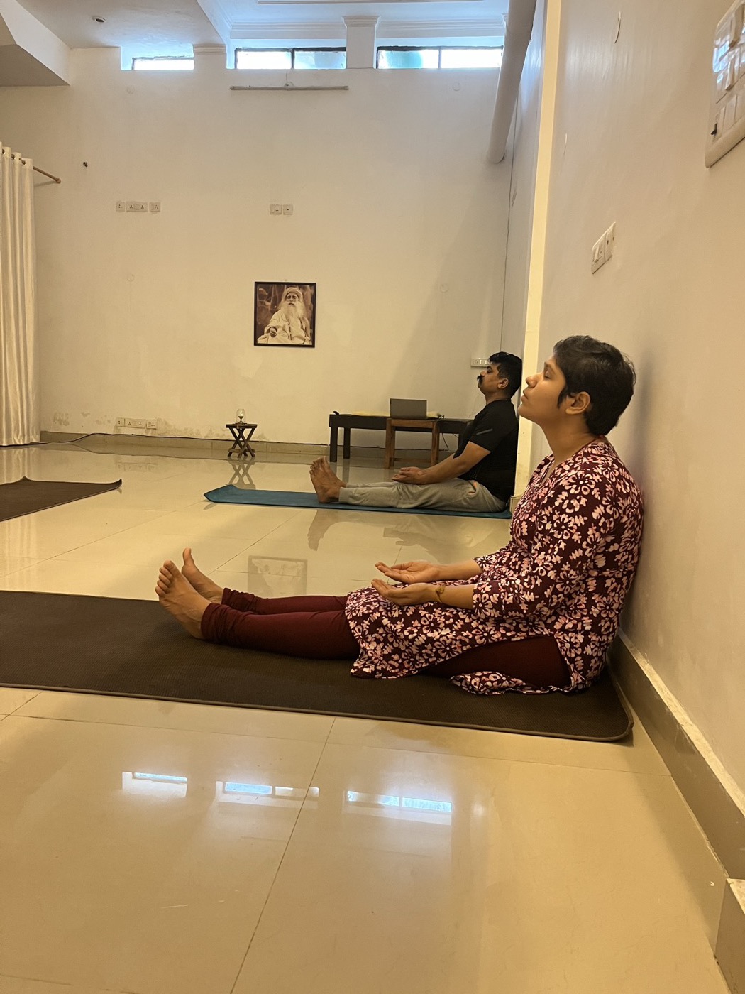 Practitioners seated upright with wall support practicing classical posture under Sadhguru portrait in Sonepat studio